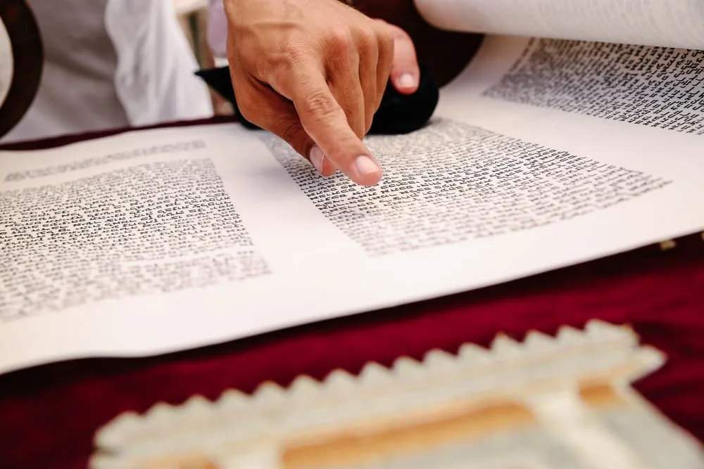 A hand tracing the Hebrew text of an open scroll