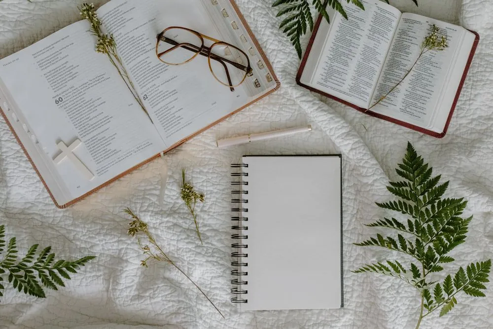Two open Bibles, a notebook, glasses and a pen laid out on a cloth for study